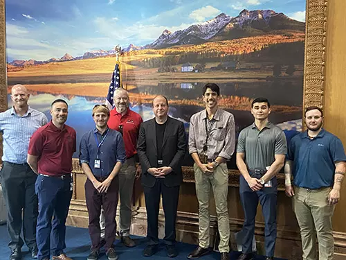 seven men of varying heights and ethnicities stand beside Gov Polis in his office in the capitol