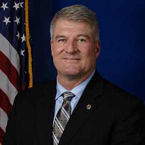 photo of stan hilkey in a dark suit and striped tie with a flag behind him