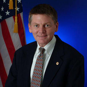 photo of kevin klein in a dark suit jacket and tie with an american flag in background