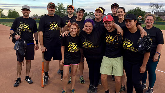 About 15 CDPS employees in black t-shirts smile on a softball field