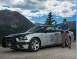 A state trooper in uniform stands next to a patrol car