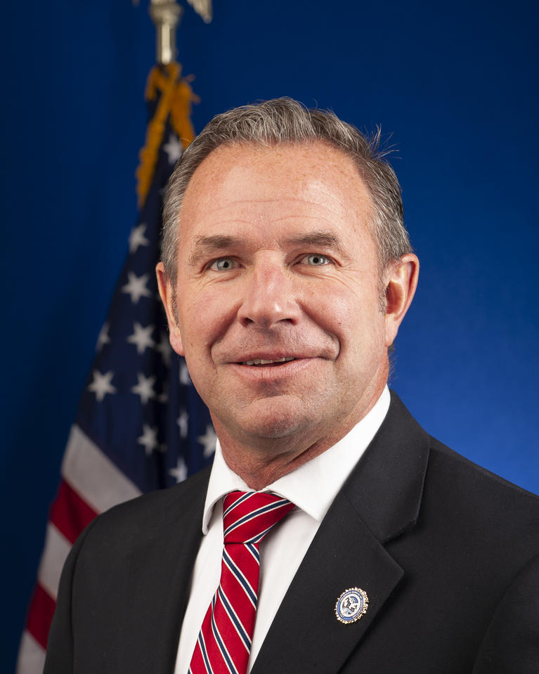 headshot of Chris Schaefer wearing a suit and tie in front of an American flag